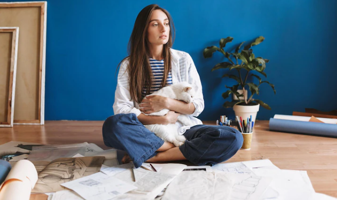 Pensive girl sitting on floor with drawings holding cute white cat in hands while thoughtfully looking aside at home